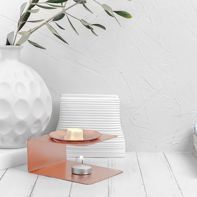 A Copper Wax Melter with a tealight candle burning underneath made by Empire Copper sitting atop a white wooden board with a white background and a small stack of books partially in the frame to the right.