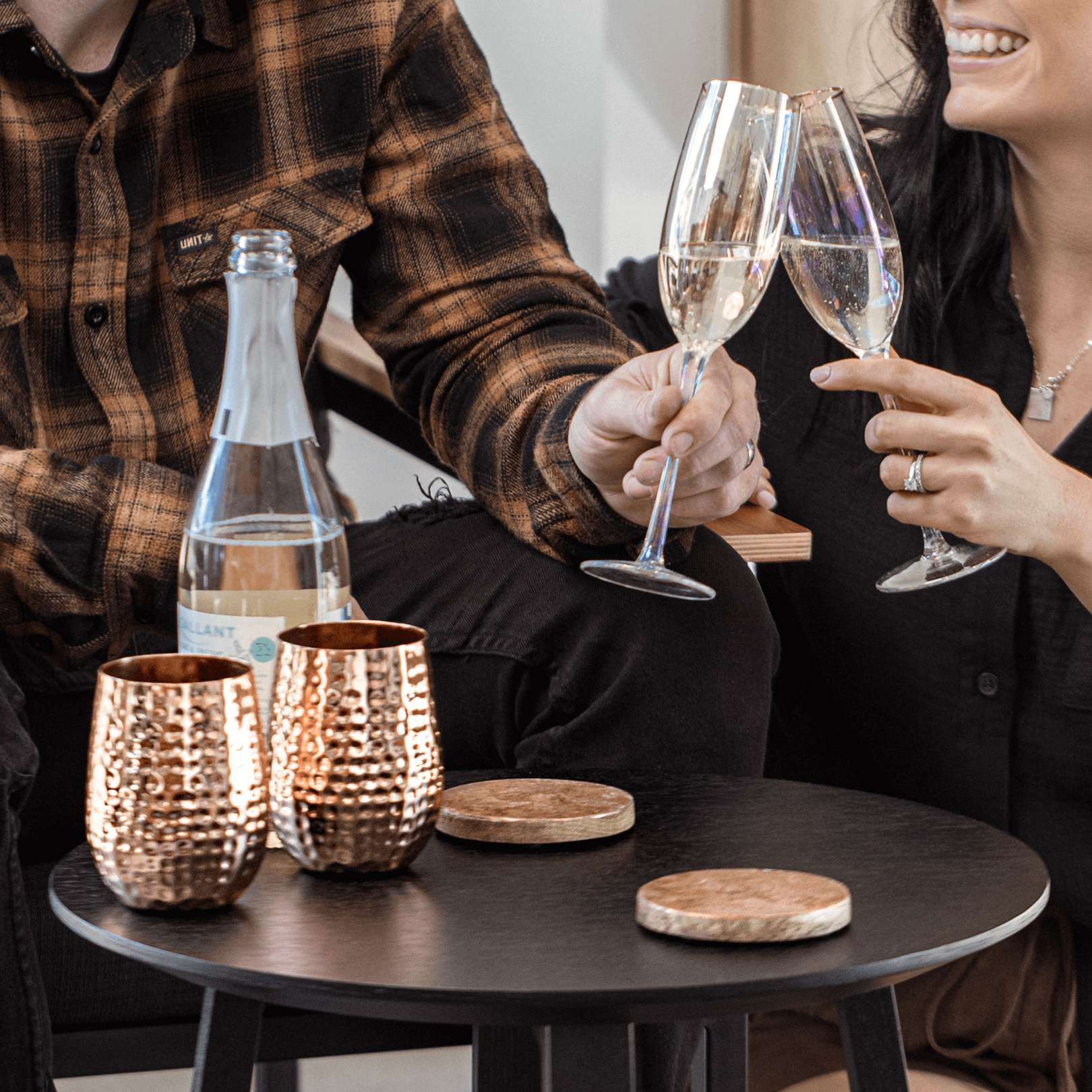 Cam and Beth Hayes cheers with two champagne flutes. There is a black coffee table in front of them. There are two copper wine glasses and two copper coasters atop the coffee table.