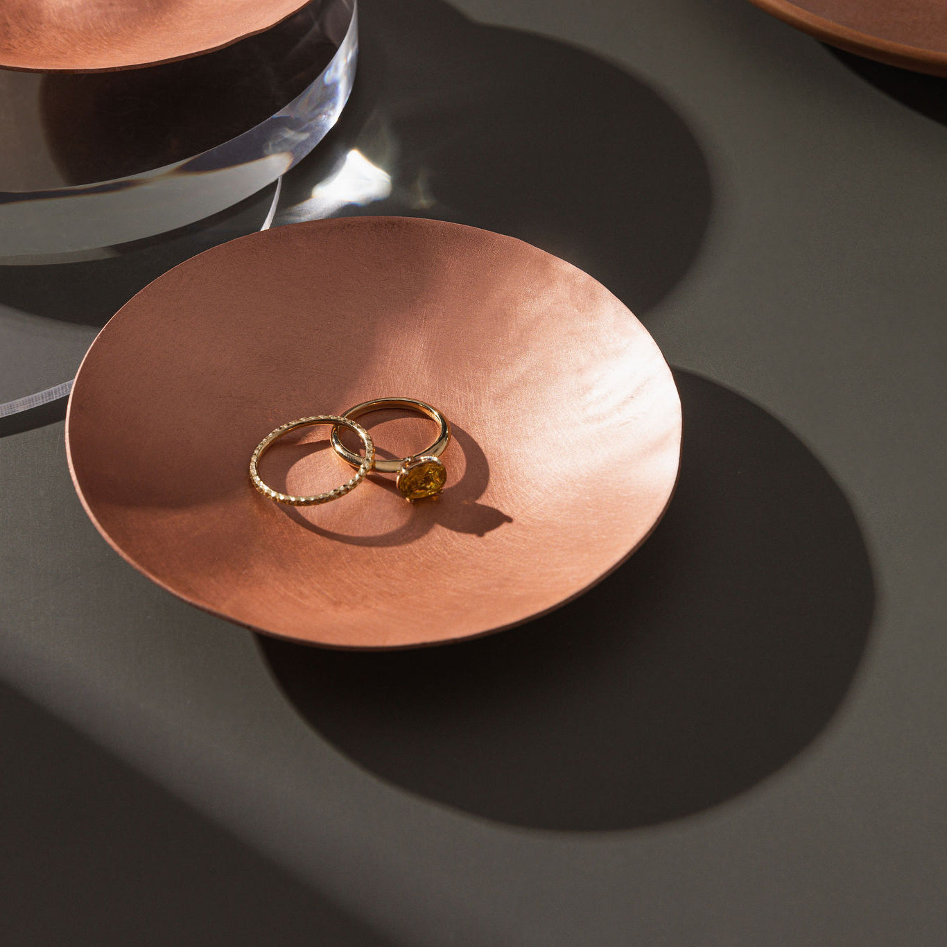 A small, round, matte copper trinket dish photographed atop a moss green background. Within in the trinket dish there is two gold rings, one with a large amber stone. The trinket dish is made by Empire Copper.
