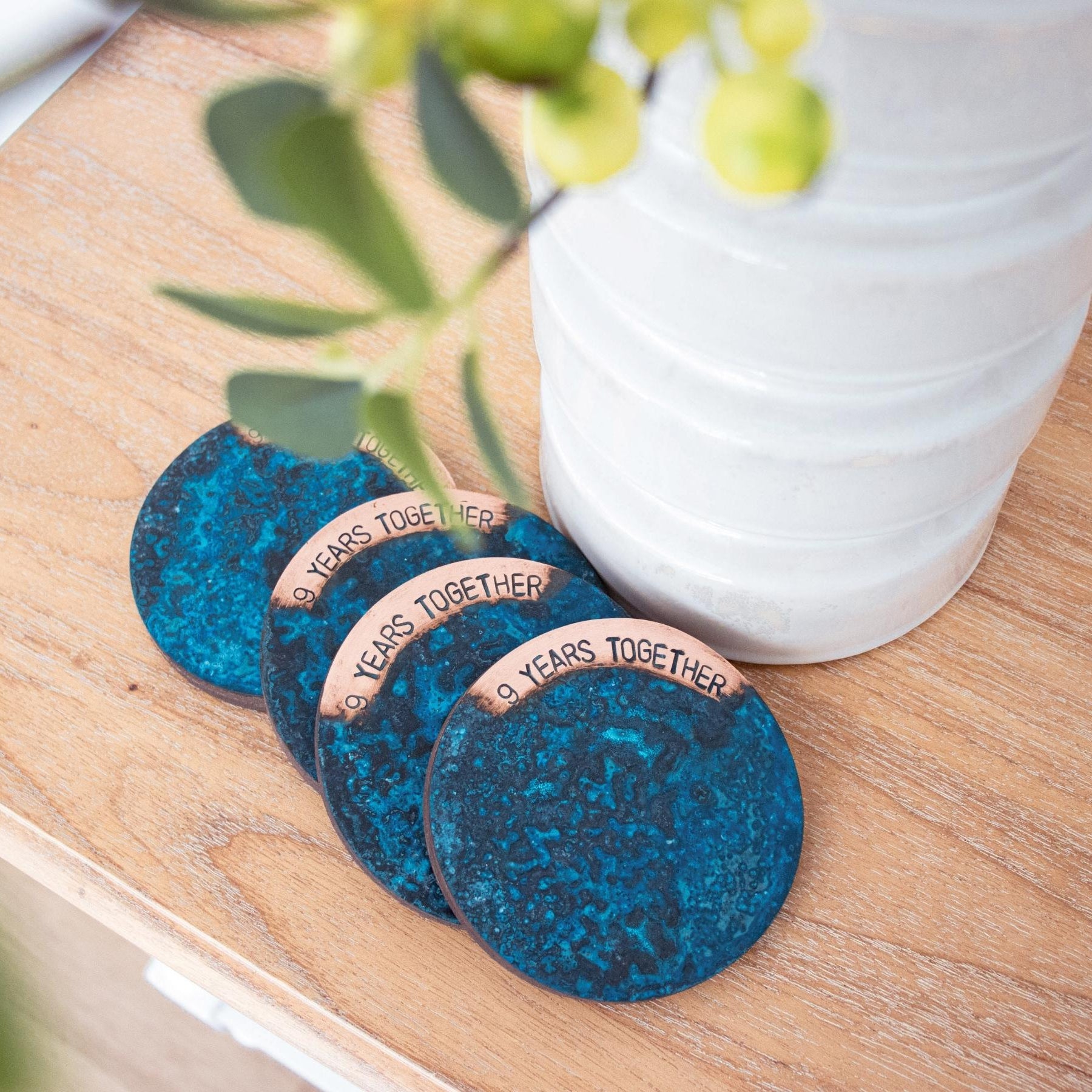A Set of 4, Blue Patina Coasters engraved with '9 Years Together'. The coaster set is made in Perth by Empire Copper and is pictured sitting atop a light oak, wooden buffet and placed next to a tall white vase.