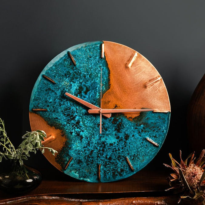 A copper wall clock with a vibrant, blue patina sits atop a wooden bench and in front of navy a background. The clock is made by the company Copper Brass and Bronze and Empire Copper by Hayes Home.
