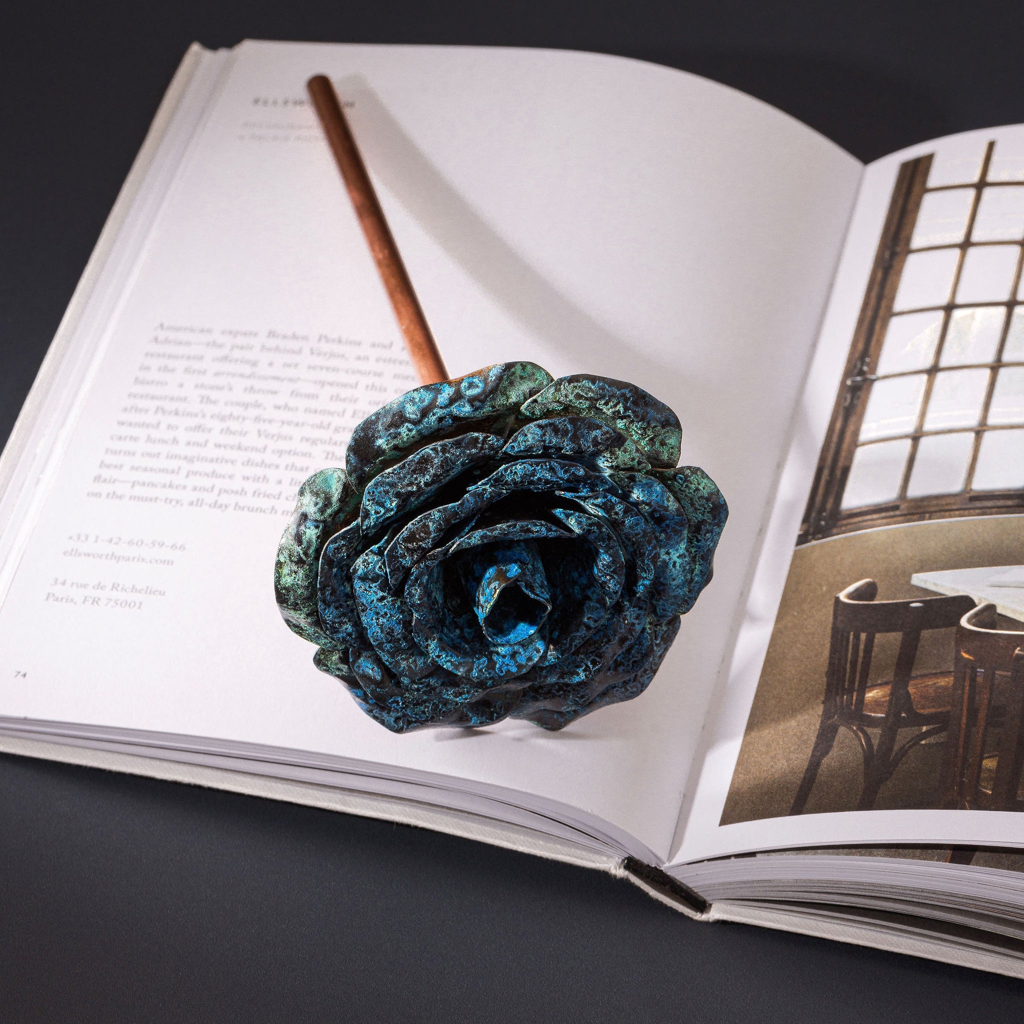 A rose made with a copper stem and a blue patina, copper rose head; lays flat across an open book. The open book is placed upon a dark navy surface. The copper rose is made by Empire Copper.