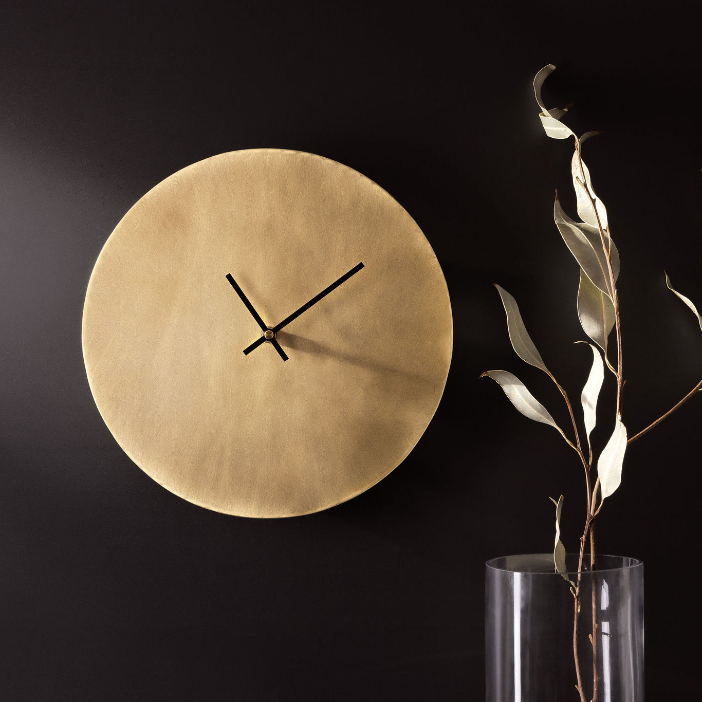 A golden, brass clock with thin black hands affixed to a dark, charcoal coloured wall. To the right of the clock is a glass vase with some long, green leaves. The picture is landscape and clock is made by Empire Copper.