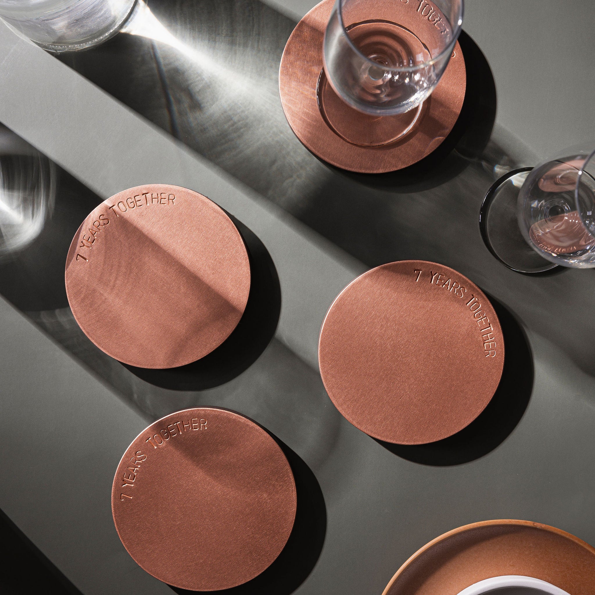 A set of four, copper coasters laying atop a moss green surface. There is a champagne flute atop one of the coasters and shadows draping across the image. This photo is taken as a birdesye view. The coasters are made by Empire Copper.