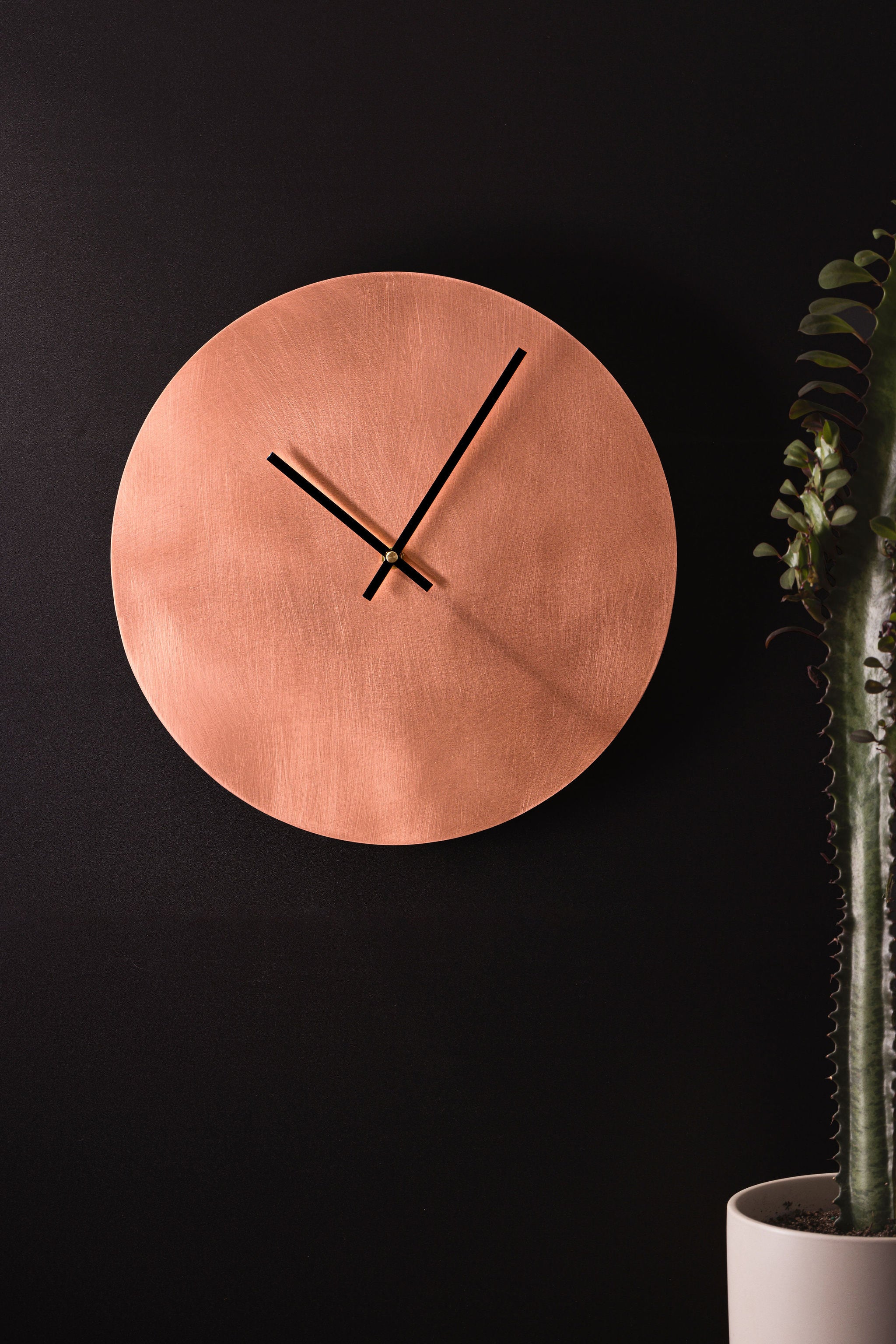 A round, glowing copper clock with thin black hands. The clock is affixed to a dark, charcoal background. To the right of the clock is a long, green cactus that is nestled in a small grey pot. The clock is made by Empire Copper.