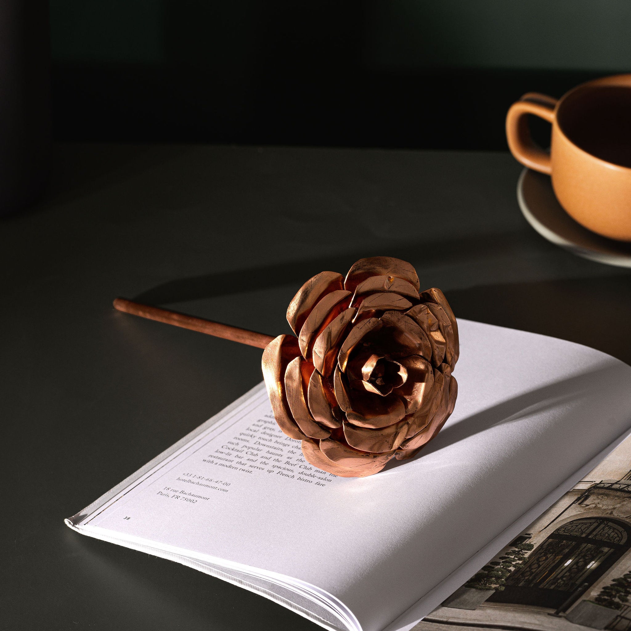 A rose made entirely from copper, laying across an open, white page book. In the background to the right, you can partially see a brown teacup and saucer. They items are sitting atop a moss green table. The copper rose is made by Empire Copper.