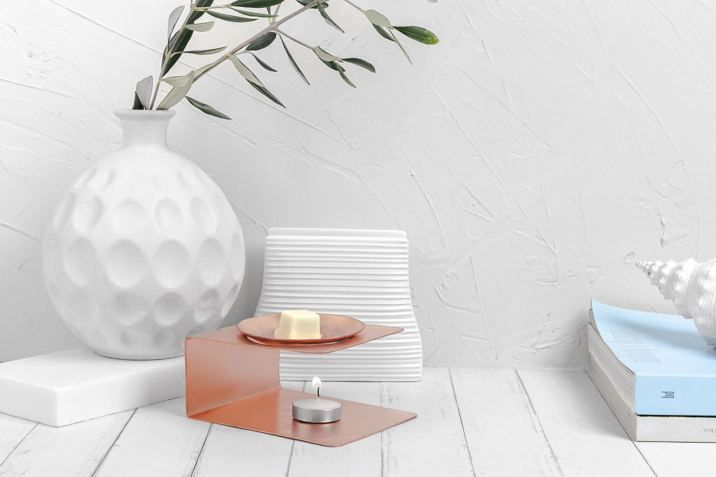A Copper Wax Melter with a tealight candle burning underneath made by Empire Copper sitting atop a white wooden board with a white background and a small stack of books partially in the frame to the right.