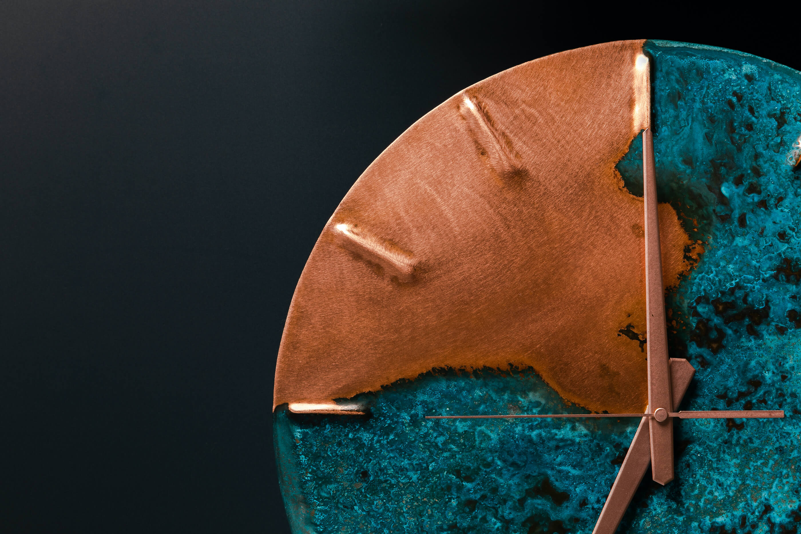 A rectangle picture with a dark navy background. On the right hand side of the picture, a copper and blue patina clock sits but it is only half in the picture. The hour hand is at number 12. The clock is made by Empire Copper.