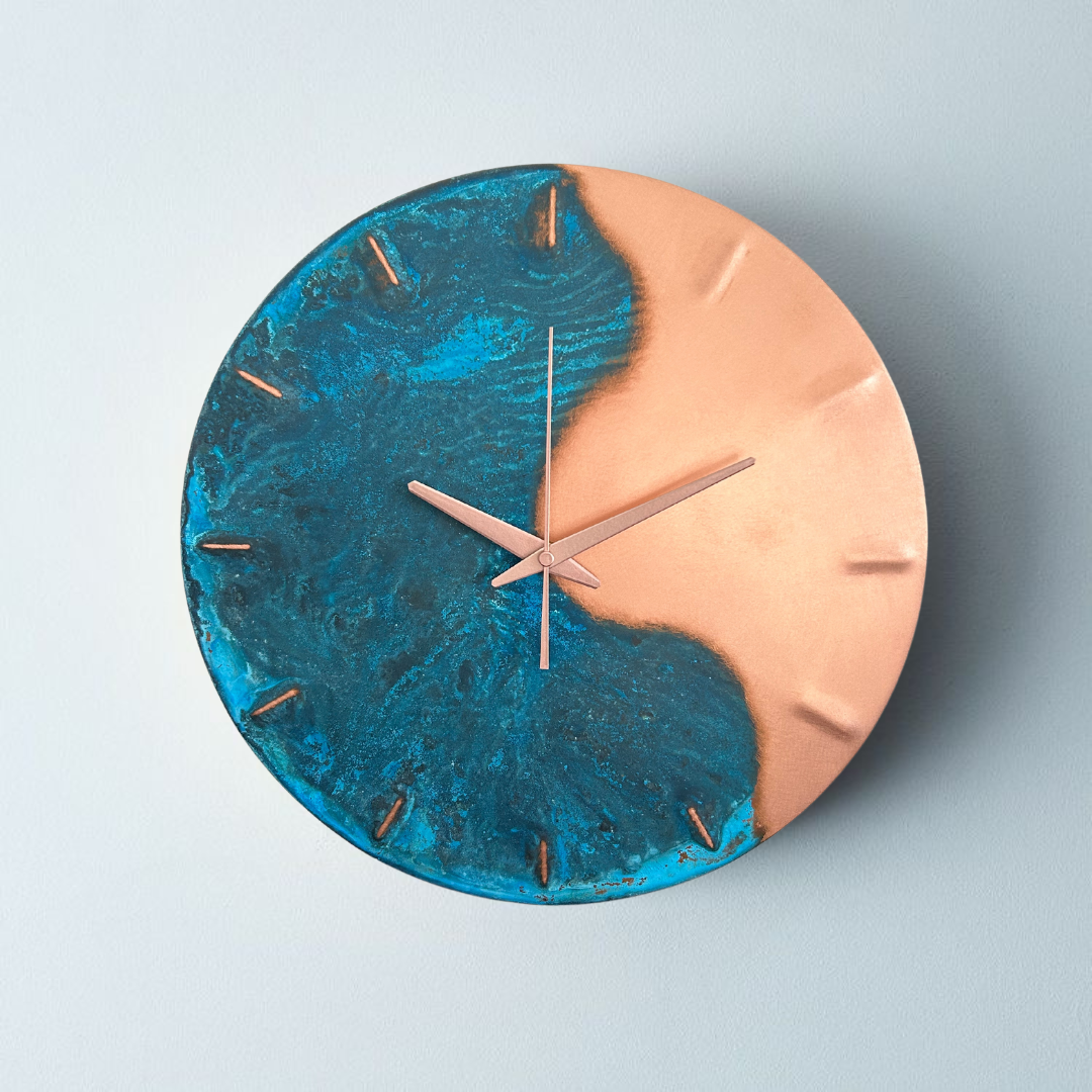 A round, blue patina, copper “half time” wall clock made by Empire Copper on a light grey background. 