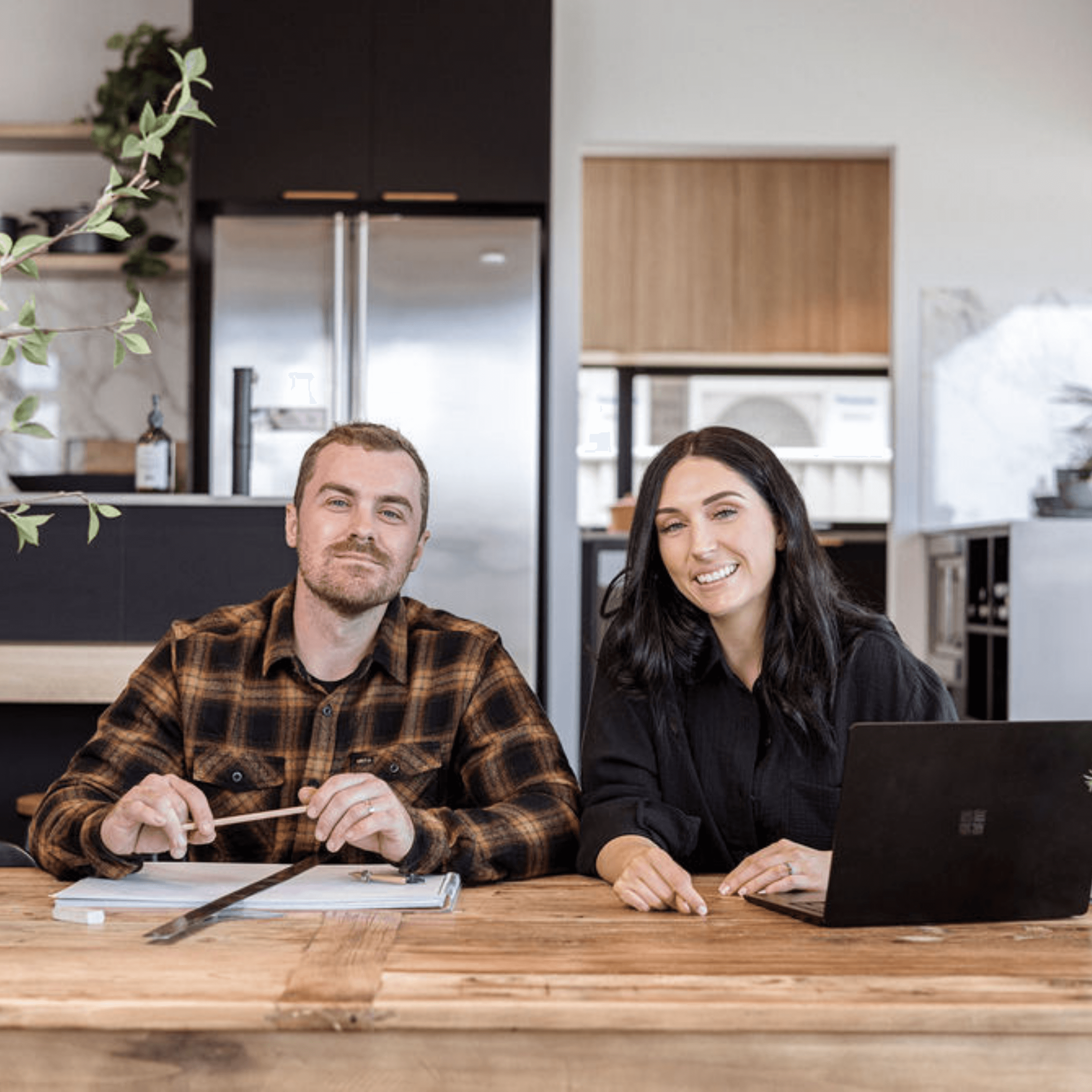Cam and Beth Hayes sitting at a wooden table in a landscape style photo. Cam is wearing an orange and black flannelette shirt and Beth is wearing a plain black shirt. They are both looking directly at the camera smiling.